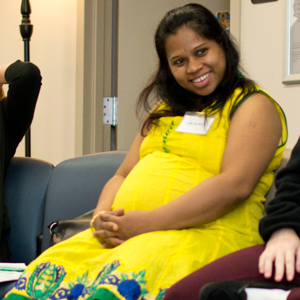Pregnant person seated in a waiting room wearing a yellow dress with green and blue floral patterns, hands clasped on lap, with office chairs and a door in the background.